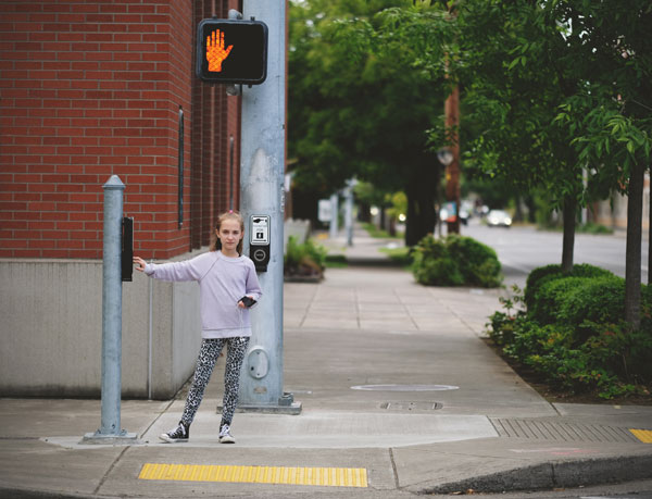 Photo of young girl pressing a crosswalk button and waiting for the signal to change at a crosswalk.