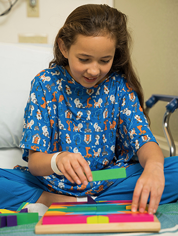 A little girl in a hospital bed plays with a toy that involves a board with blocks of different shapes.