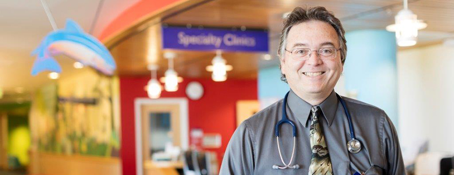 A doctor smiling in a clinic lobby with a sign reading "Specialty Clinics" in the background.