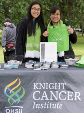 Two women smiling while standing behind a table with the "OHSU Knight Cancer Institute" logo on the front.