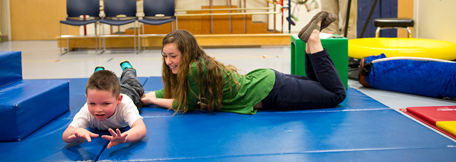 A female physical therapist guiding a young boy through physical therapy exercises on a mat.