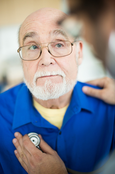 An elderly patient watching as a doctor listens to his heart with a stethoscope.