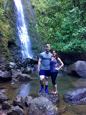 A man with his arm around a woman pose in front of a waterfall while on a hike.