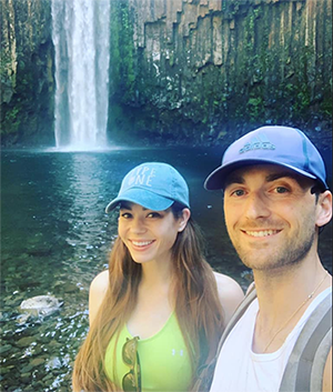 A man and a woman smiling taking a selfie with a waterfall in the background.