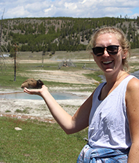 A woman posing with her hand out to give the impression that she is holding a buffalo that is sitting down off in the distant background.