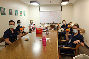A group of pediatric residents sitting around a conference table wearing their scrubs and PPE masks, each of them holding up an orange.