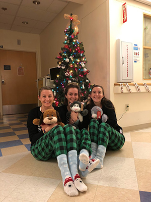 Three female pediatric residents sitting on the ground holding stuffed animals in front of a holiday tree at an OHSU clinic.