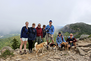 Four women, three men and three dogs in a line posing while on a hike on a cloudy day.