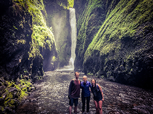 Two women and one man stand in a swimming hole with a waterfall behind them.