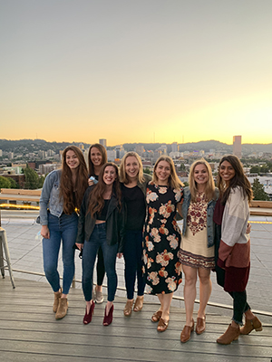 A group of women pose in a line on a rooftop bar at sunset with downtown Portland behind them.