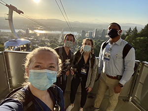 Four OHSU Pediatric Residents wearing PPE masks (two women and two men) standing at the OHSU Tram platform on a sunny morning.