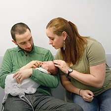 Photo of a man sitting down holding a newborn baby and a woman sitting next to him leaning over touching the baby's head.