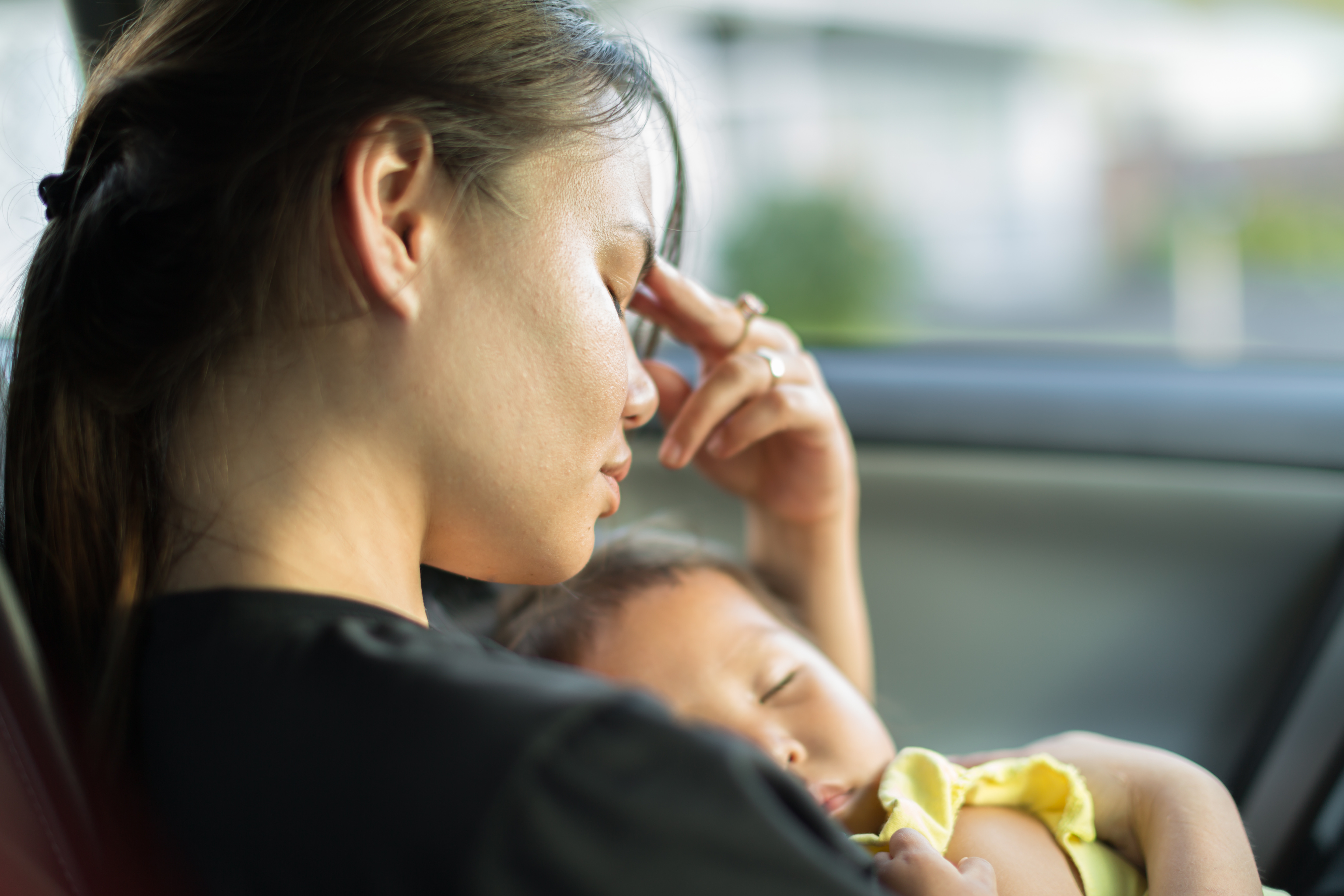Woman sitting in car holding sleeping baby. She looks tired and anxious.