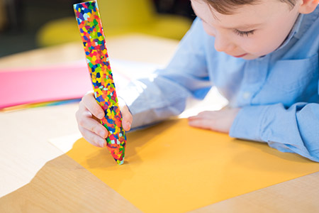 Photo of a child coloring on yellow paper with an oversized multicolor crayon.