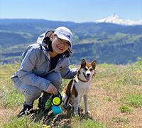 A woman crouching down smiling next to her dog while on a hike with Mt. Hood in the background.