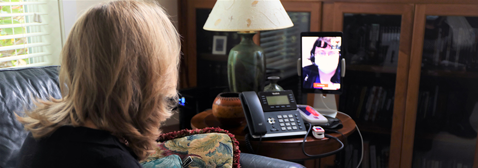 A woman looking at a side table that has a telephone as well as a propped up tablet that is being used to videoconference with a healthcare provider.