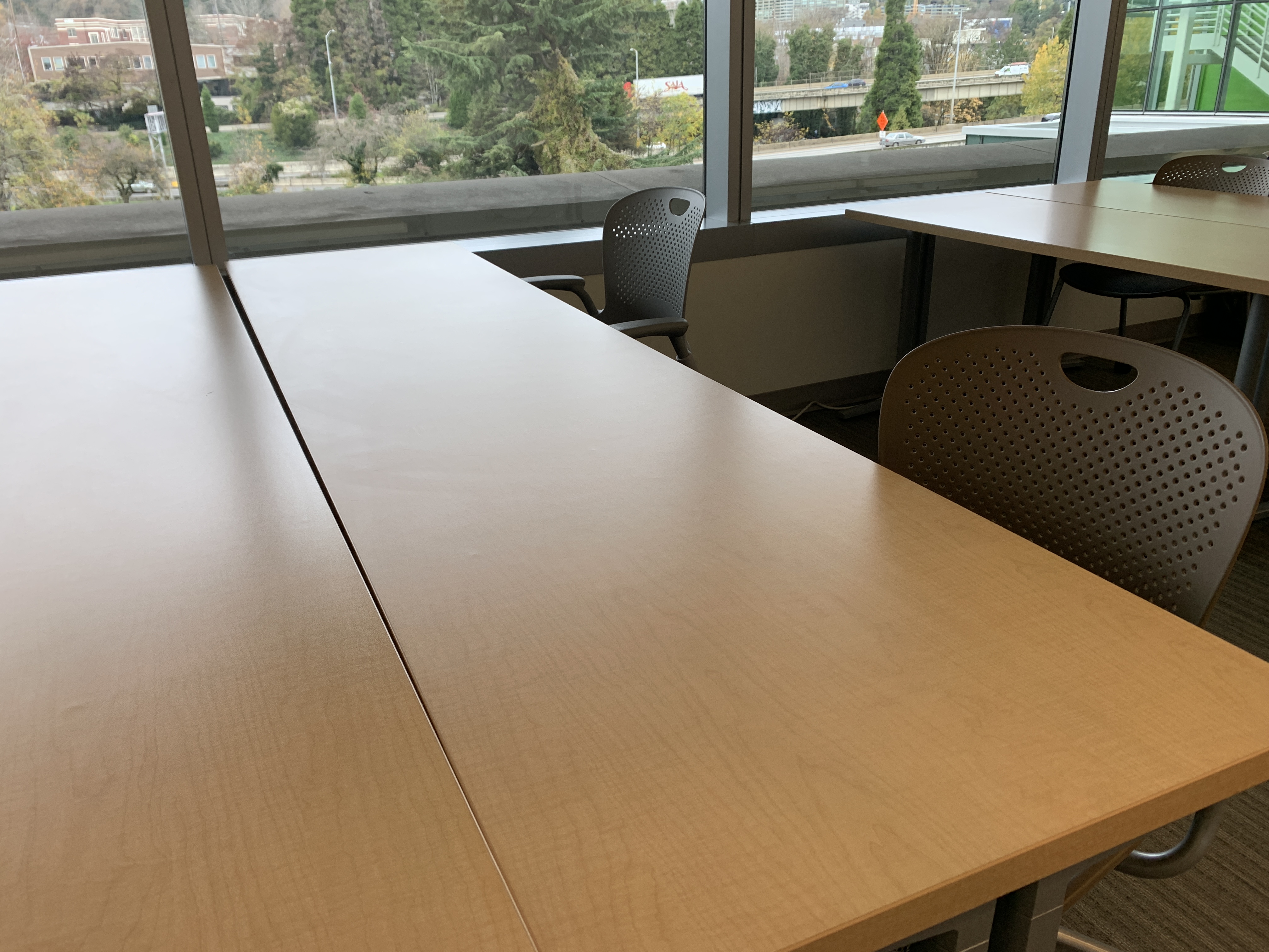 A large work table in a library space with two chairs pulled up. In the background is a wall of windows.
