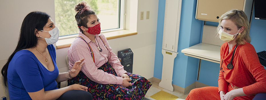 Two women sitting in a hospital exam room speaking to a female healthcare provider, also sitting. All three are wearing PPE masks.