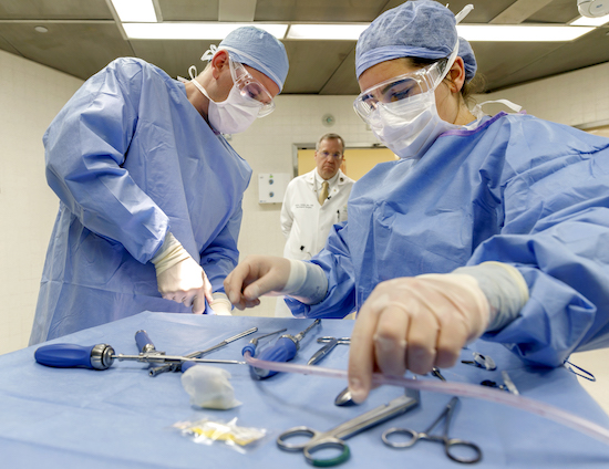 Two students in scrubs, gloves, and masks arrange surgical equipment on a tray.