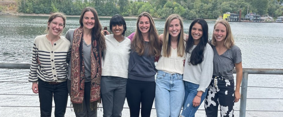 A group photo of the seven women comprising the 2025 OHSU OB/GYN Residency Class standing against a railing in front of a lake.