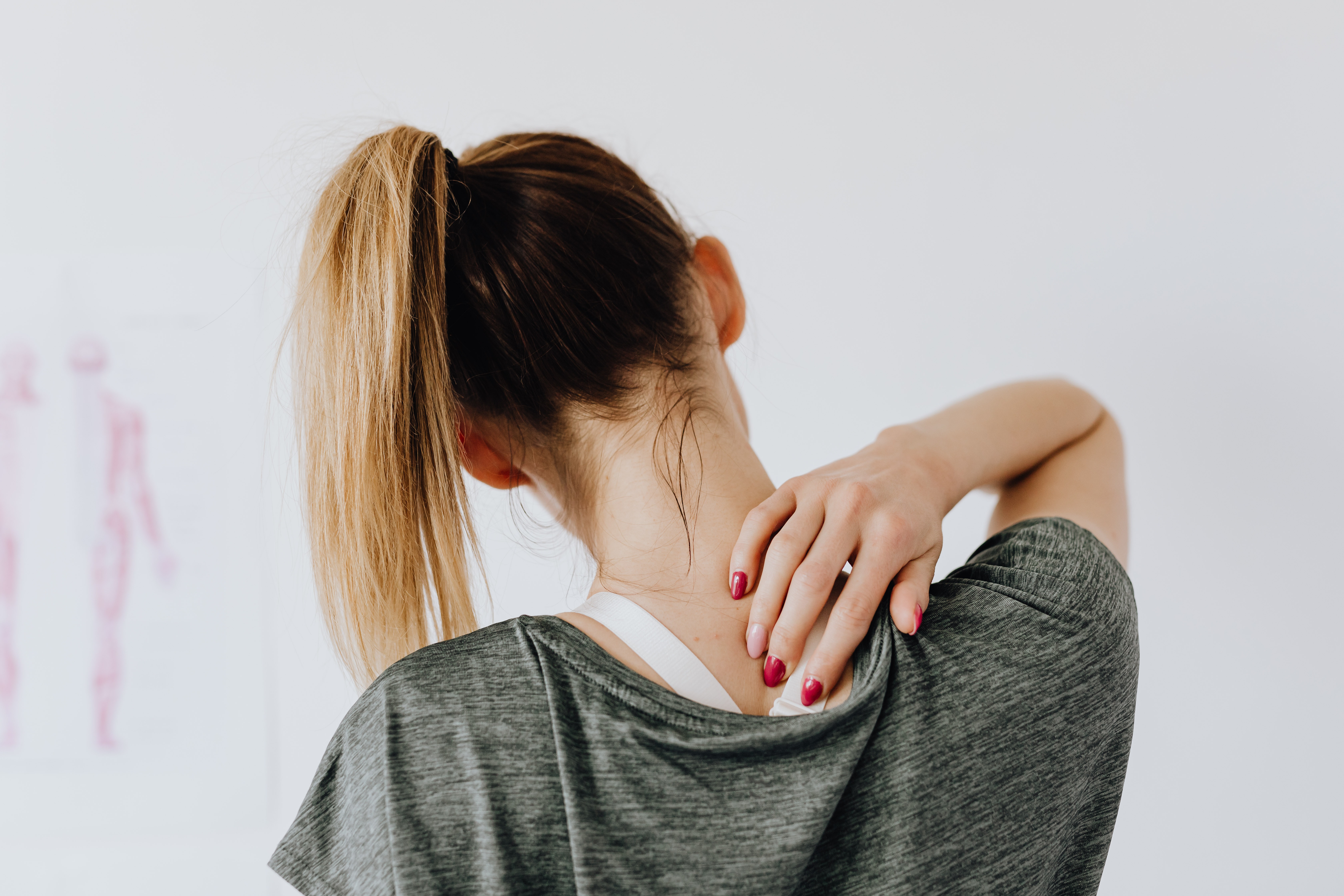 A teen girl's head and shoulders, viewed from the back as she indicates neck or back pain. The girl has dyed blonde hair and red painted fingernails.
