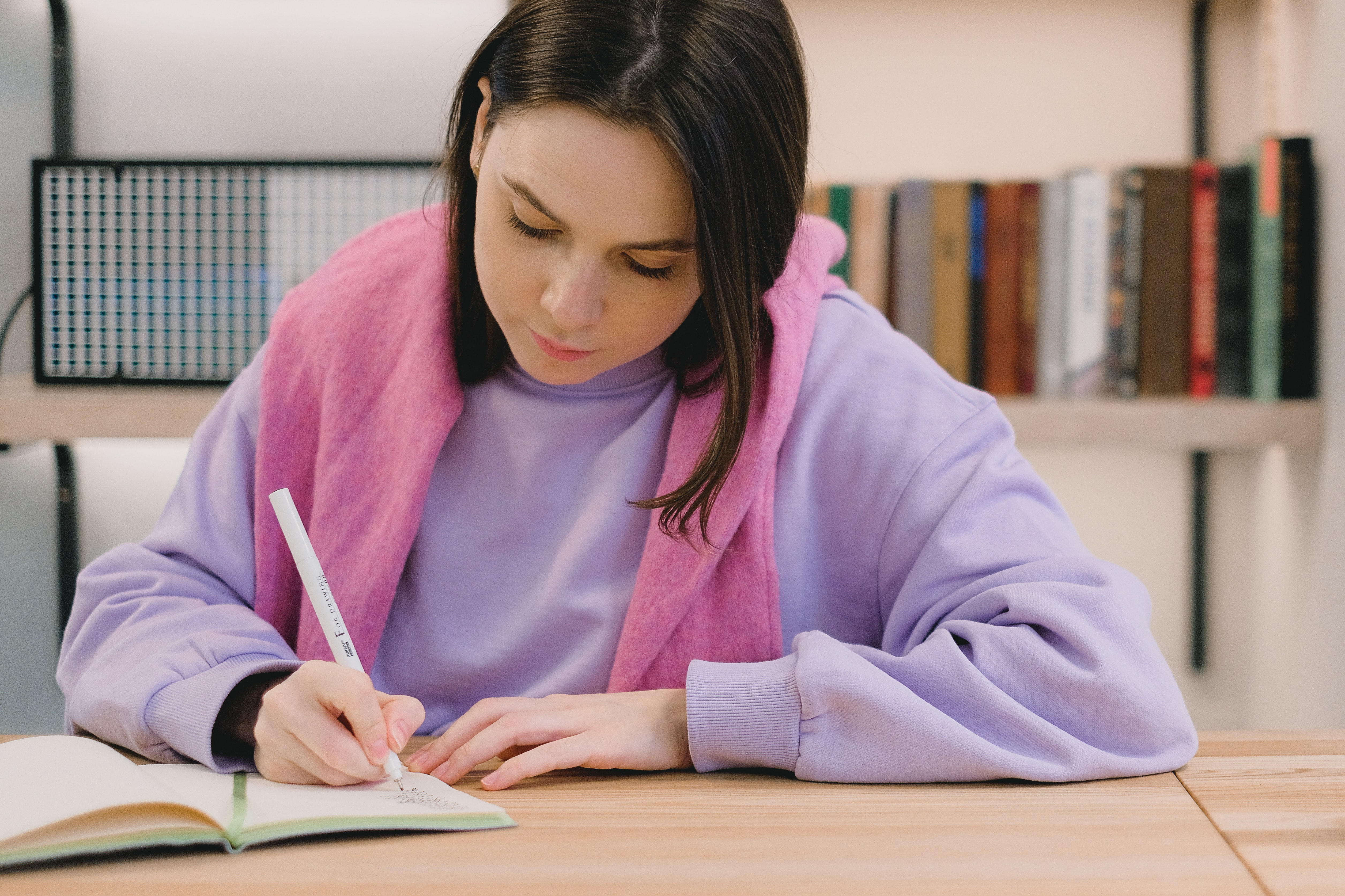 A teenaged girl with dark hair and light skin studiously writes in a notebook. She is wearing a light purple sweater and pink scarf.