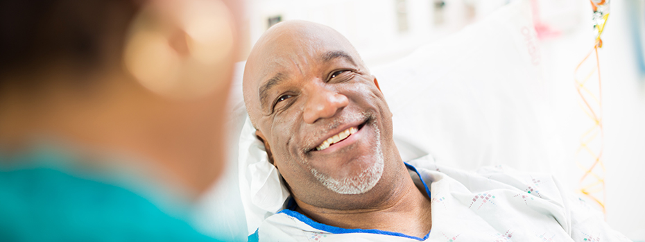 A patient lying in a hospital bed looks up smiling at a provider who is at his bedside.
