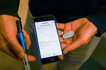 Hands holding glucose sensor transmitter, a smart insulin pen and phone displaying DailyDose app.