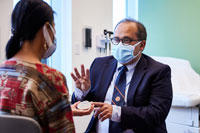 A female patient listening to a male doctor as he provides an explanation in an exam room, both sitting and both wearing PPE masks.