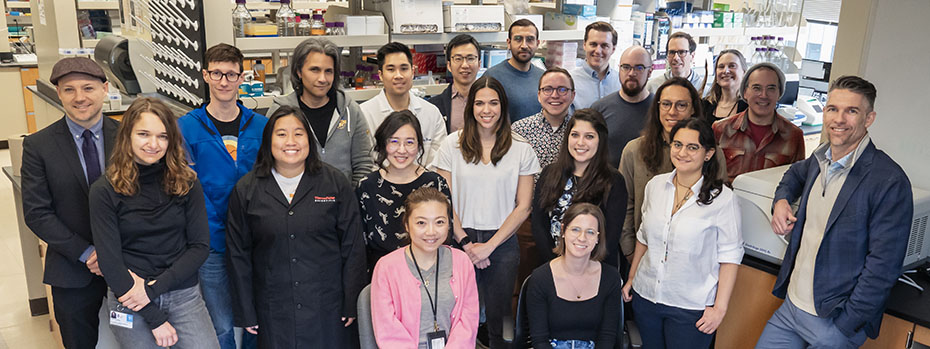 A large group of people, including students and faculty, gather in the biomedical engineering lab for a photo and smile at the camera.