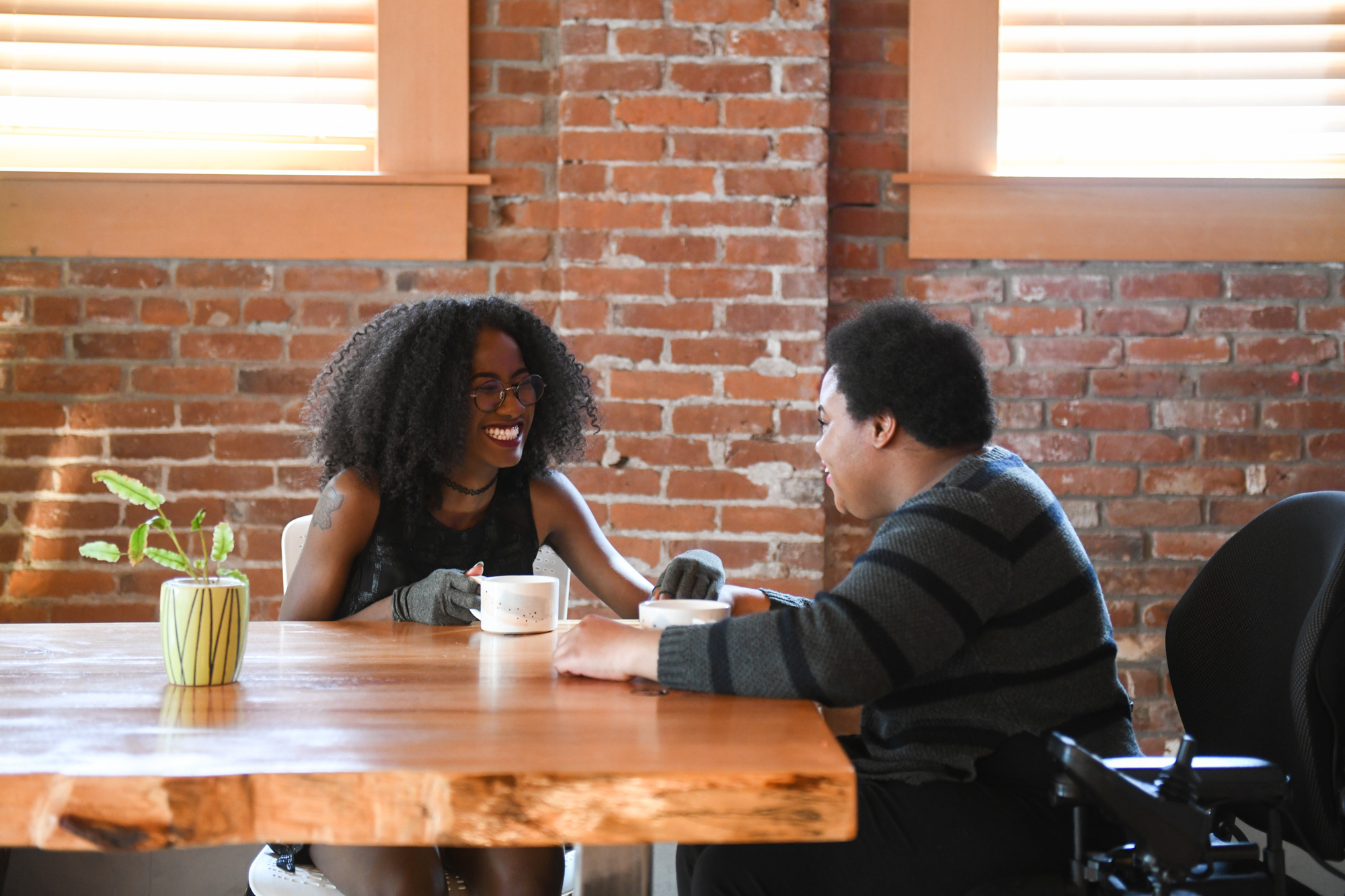 Two disabled Black people (a femme wearing compression gloves and a non-binary person in a power wheelchair that's partially in view) sit across each other and laugh while on a coffee date in a brick building.