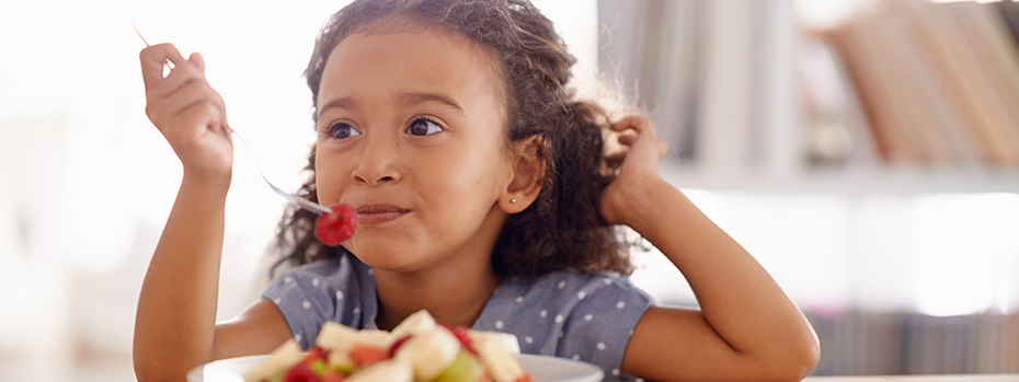 A girl holds a fork with a strawberry over a dish of fruit salad.