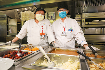 Two OHSU employees wearing face masks stand behind a hot food station with mashed potatoes, gravy and other choices.