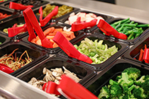 Multiple pairs of plastic tongs are ready to use at a salad bar offering broccoli, mushrooms, celery, green beans, carrots and more.