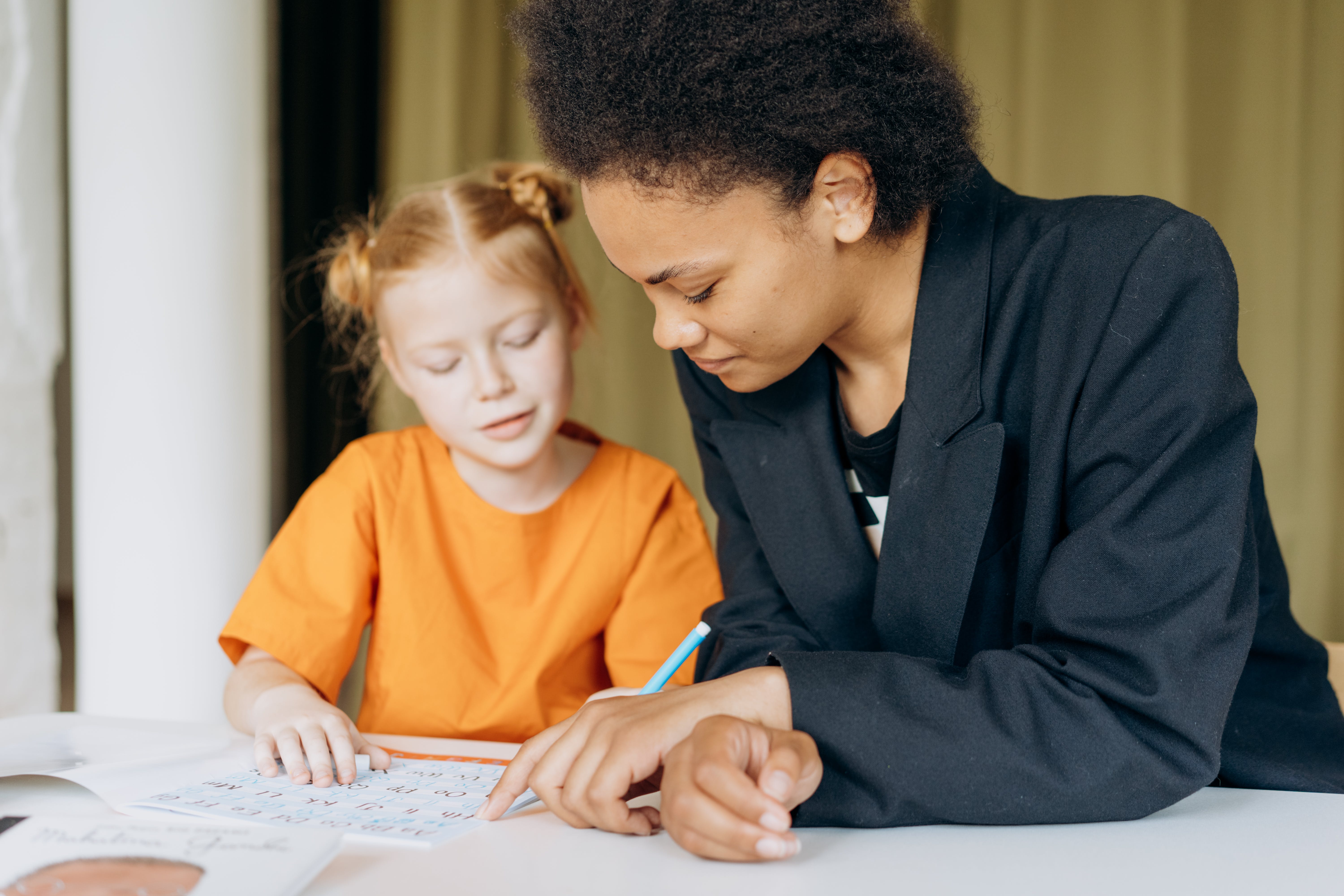 A young girl writing on a workbook sits next to a woman in a blazer.