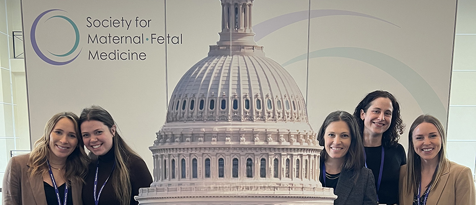 Five women standing in front of a sign that reads "Society for Maternal-Fetal Medicine," with an image of the U.S. Capitol Building in between them.