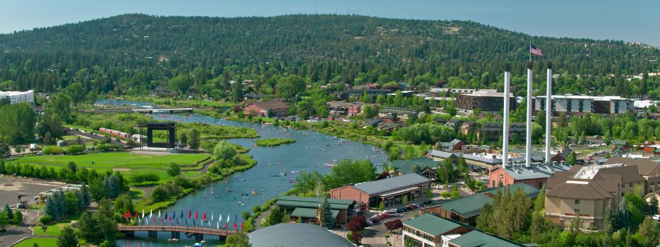 An aerial shot of the town of Bend, Oregon. It includes the Deschutes River, the Old Mill, and mountains in the background on a sunny day.