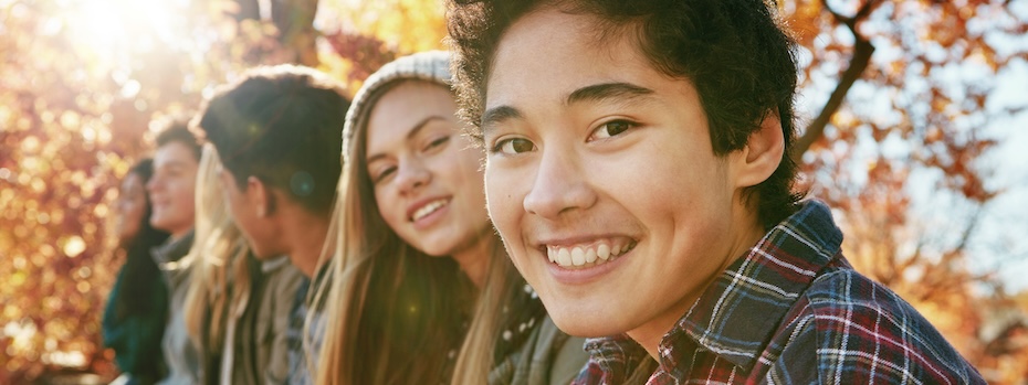 A youth smiles while sitting outside with other young people on a sunny day near trees with fall foliage.