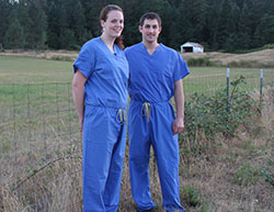 Two people in blue scrubs pose in front of a grass field