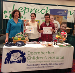 A photo of two women and a man standing behind a vendor table that has a cover that reads "Doernbecher Children's Hospital."