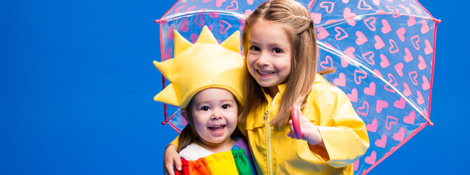 A little girl in raincoat holding an umbrella, with her arm around a toddler, who is dressed in a "sun" headdress and a "rainbow" jumper.
