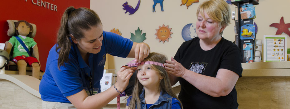 A young girl patiently waiting as two women measure her head circumference with a pink measuring tape.