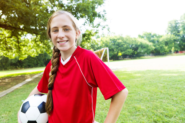 Girl her hair in a long braid, smiling and holding a soccer ball under her arm.