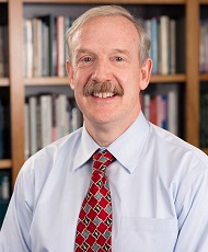 Headshot of a smiling man with gray hair and a mustache wearing a blue shirt and red tie