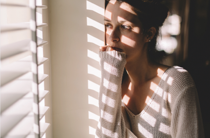 Woman stands at window with shadow of blinds on her looking thoughtful