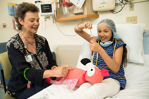 A health care provider sitting bedside next to a child patient as she pretends to give a stuffed animal its shot.