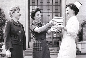 three women stand in front of the OHSU Auditorium, one hands another dressed as nurse a stack of books