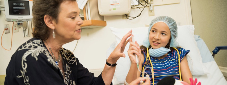 A female care provider explains an upcoming procedure to little girl who is sitting up in a hospital bed.