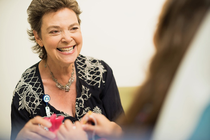 A female care provider smiles as she talks with a patient who is sitting up in a hospital bed.