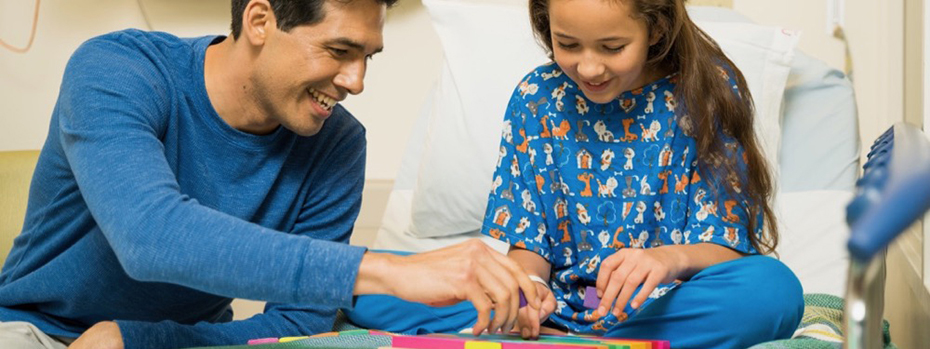 A father playing a game with his daughter as she is sitting up in a hospital bed.